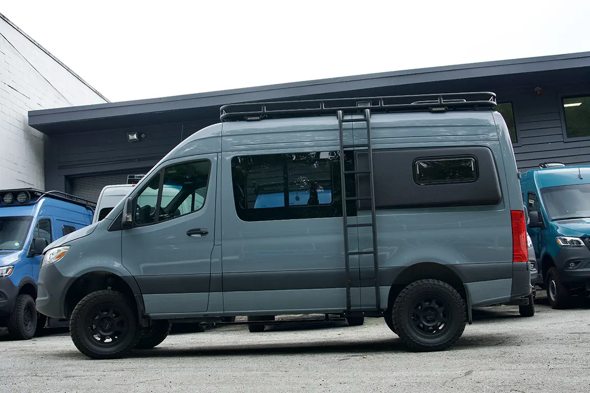 Gray Mercedes Sprinter van with roof rack and off-road tires parked outside commercial building.