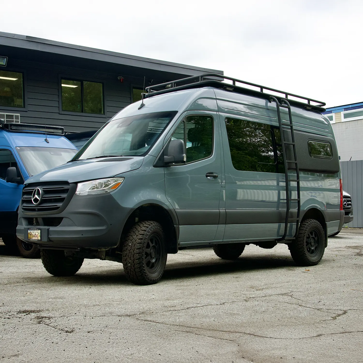 Gray Mercedes Sprinter van with roof rack and off-road tires parked outside commercial building.