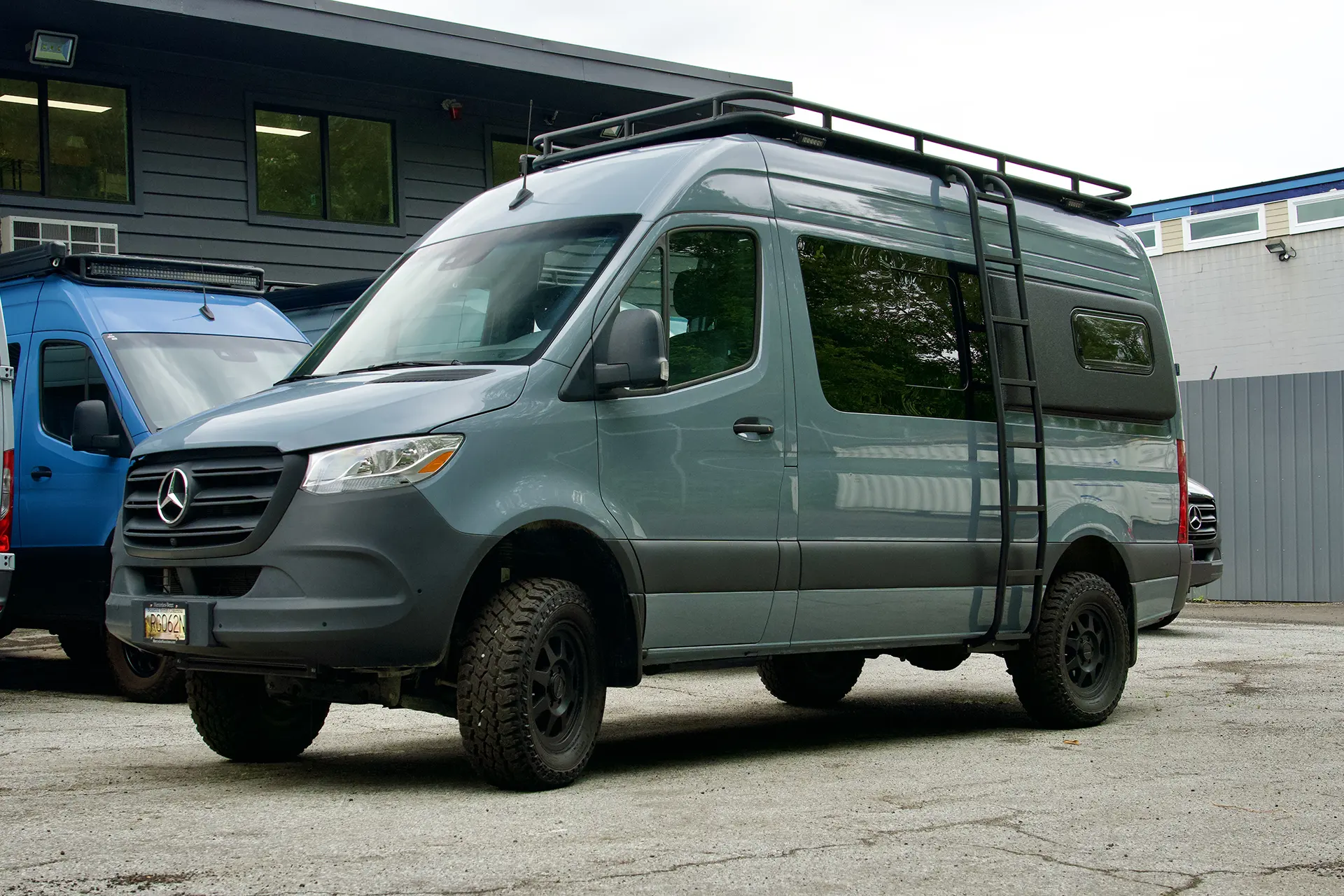 Gray Mercedes Sprinter van with roof rack and off-road tires parked outside commercial building